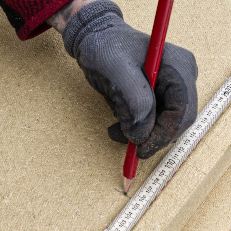 Hand im Arbeitshandschuh zeichnet mit Zimmermannsbleistift und Zollstock an